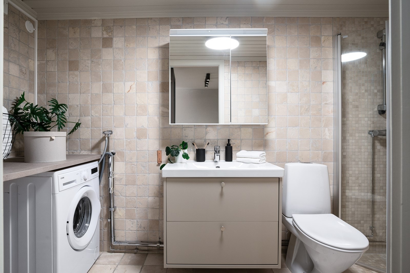 Pent og funksjonelt kombinert bad/vaskerom med fliser på gulv og vegger i lyse, nøytrale toner. Det er varme i gulvet. Galleribilde