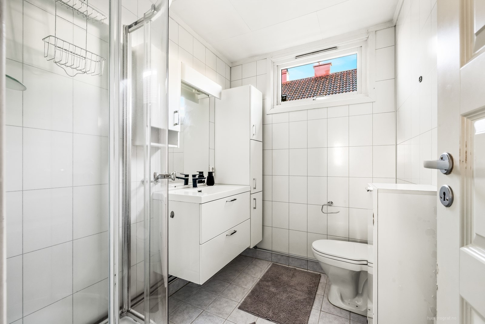 Badet har baderomsplater på veggene, flislagt gulv og takess i tak. Servant i skuffeinnrending, dusjkabinett og toalett. Galleribilde