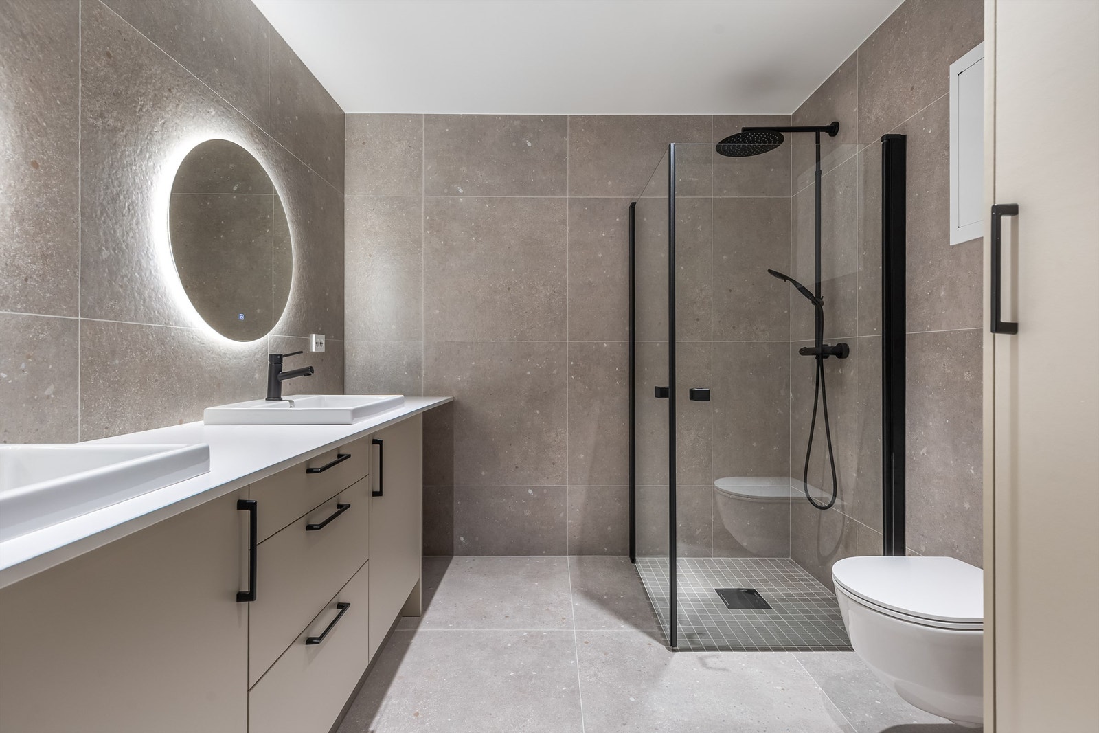 Baderom med dusjhjørne, dobbel servant, vegghengt toalett og skap med varmtvannsbereder og plass til vaskemaskin. Galleribilde