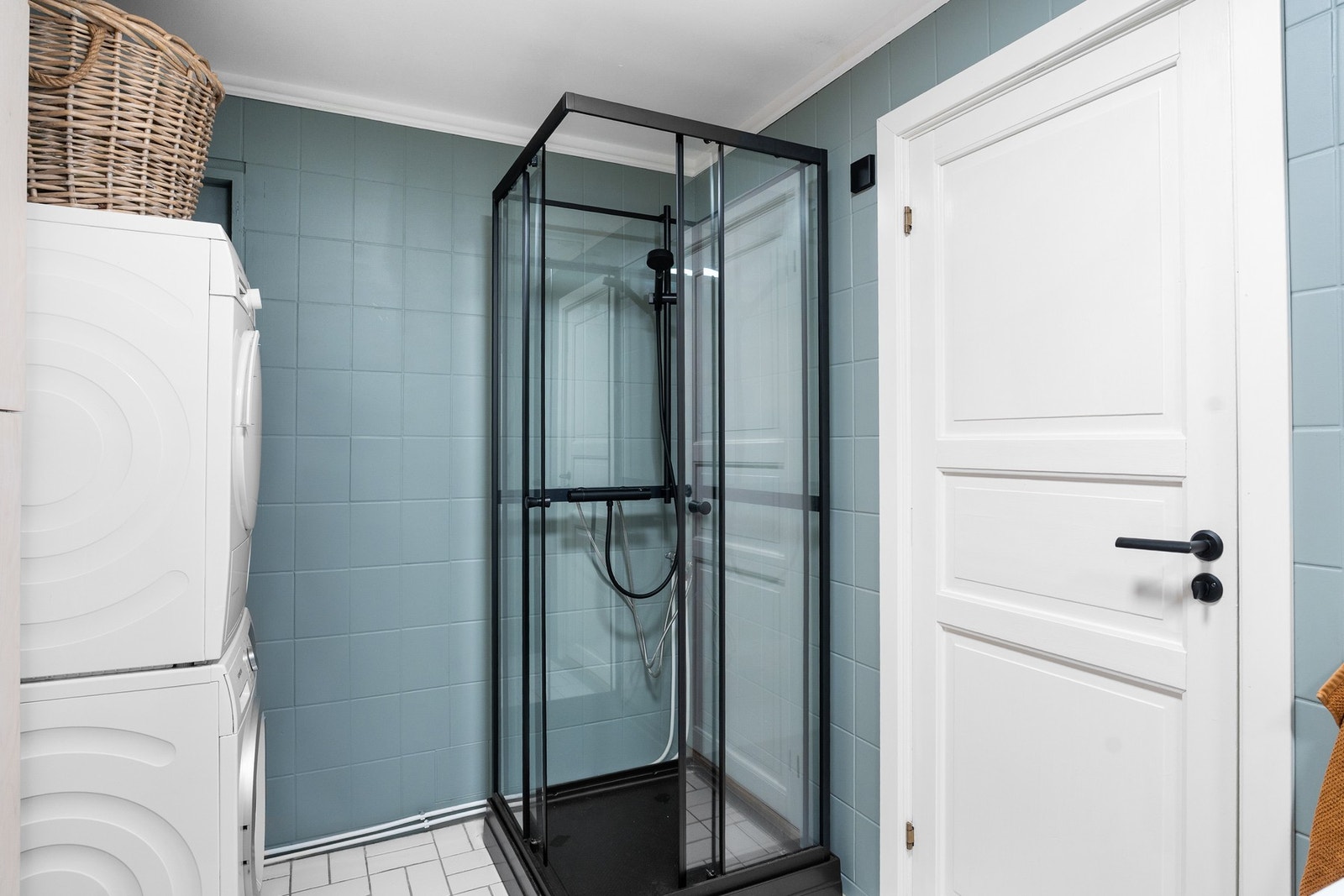 Badet inneholder servant med ettgrepskran og skuffer, rundt speil over vasken, gulvstående toalett, opplegg for vaskemaskin samt dusjkabinett Galleribilde