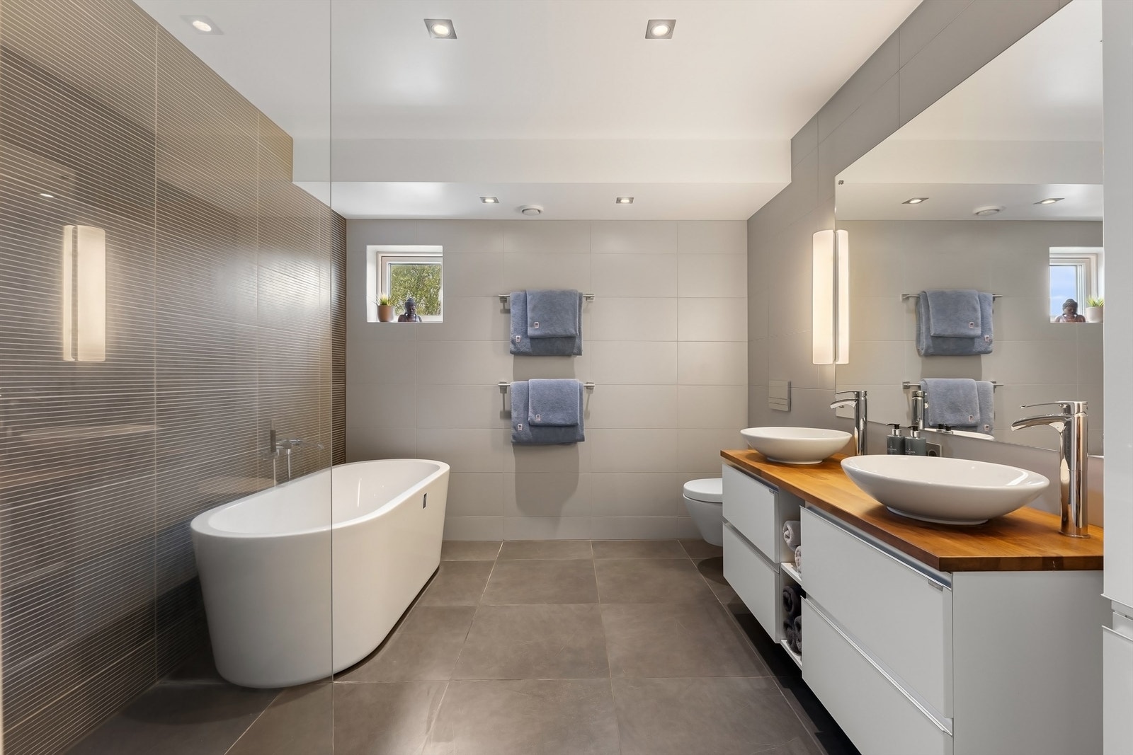 Stilrent bad fra 2014 med flislagt gulv og vegger, gulvvarme og nedsenket himling med downlights. Servantskap med trebenkeplate, to servanter, speil og belysning. Vegghengt toalett og høyskap, dusjhjørne med regn- og hånddusj, samt frittstående badekar Galleribilde