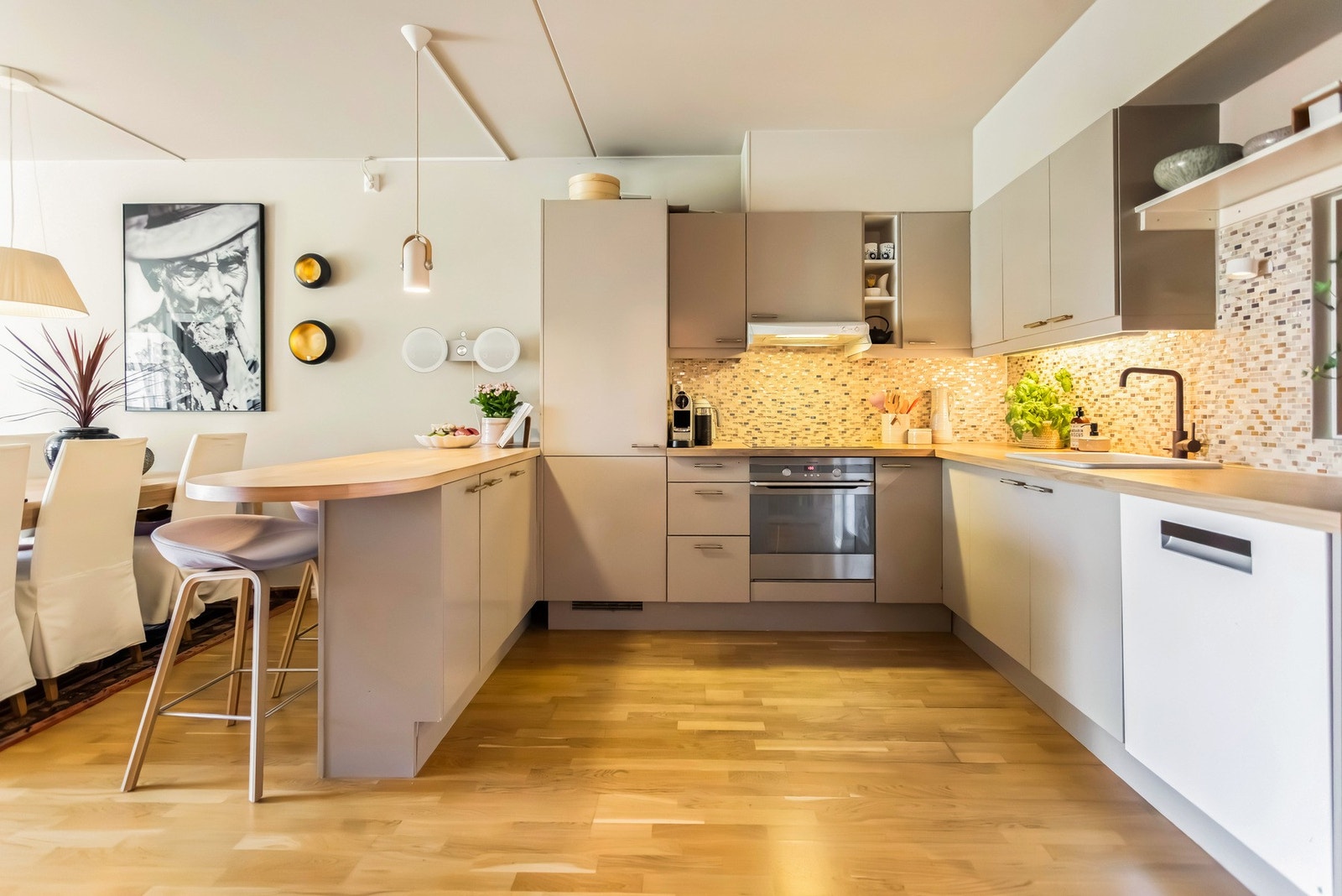 Kjøkkenet fremstår moderne og funksjonelt med slette, lakkerte fronter og heltre benkeplate som gir et tidløst uttrykk. Her finner du nedfelt porselenskum, platetopp med ventilator og stilrene fliser over benk. Galleribilde