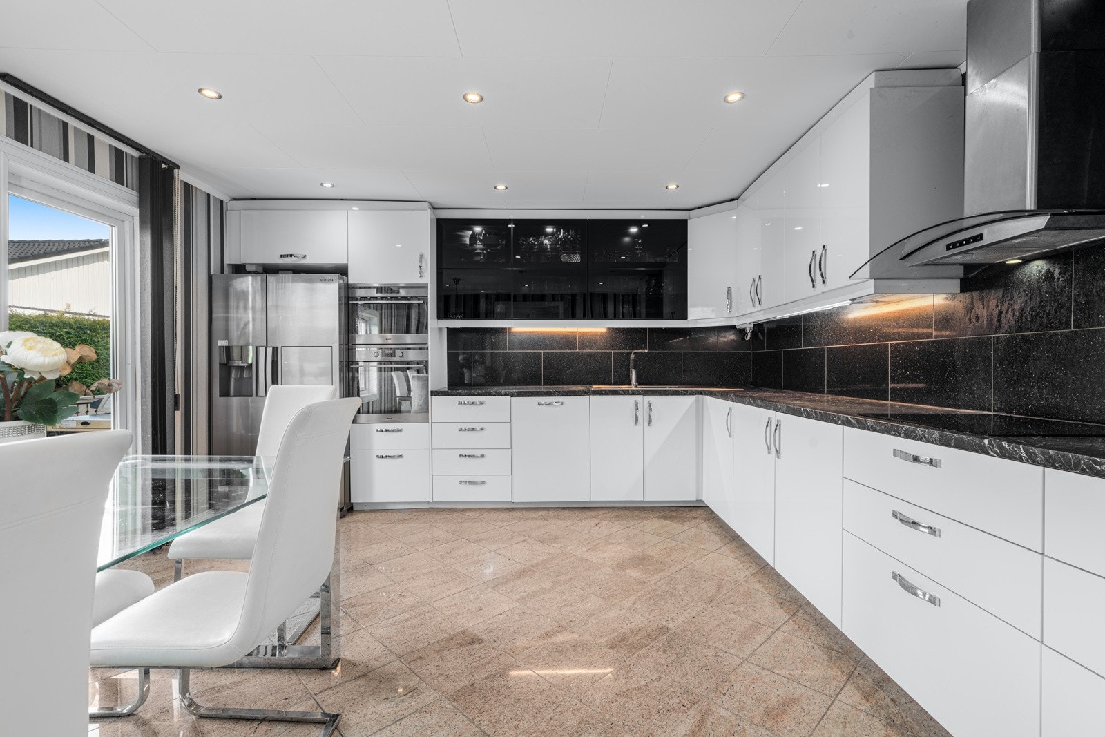 Gjennomført og moderne kjøkken med hvite fronter, svarte glassfronter og matchende svart benkeplate. Svarte fliser langs veggen binder det hele sammen og gir et stilrent uttrykk. Galleribilde