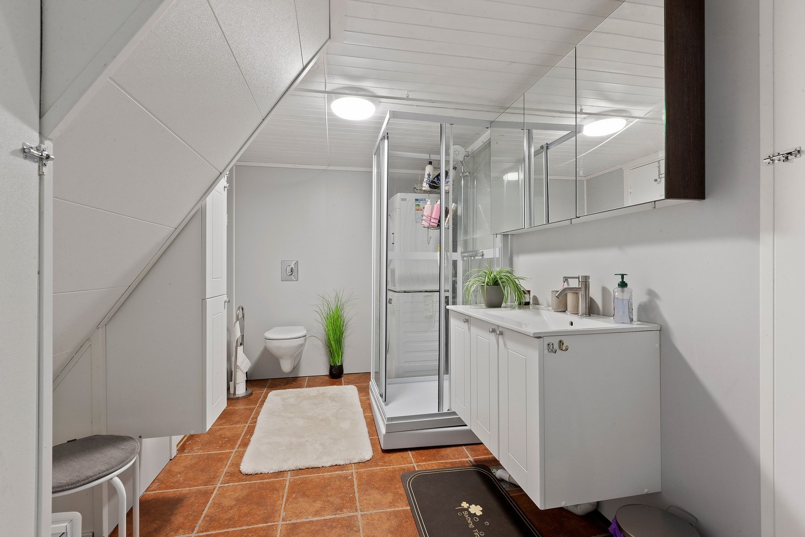 Badet har lys innredning med nedfelt servant og skap under, overskap med speil over, dusjkabinett, veggmontert toalett og opplegg for vaskemaskin. Galleribilde