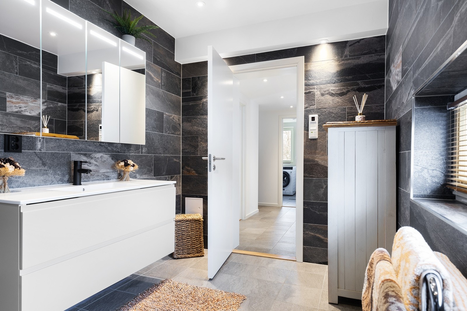 Badet i 1. etasje er flislagt på gulv og vegger, og har varme i gulvet. Veggmonert toalett og dusjhjørne. Heldekkendeservant og speil. Baderomsinnredning med laminerte fronter/skuffer, og overskap med speildører. Galleribilde