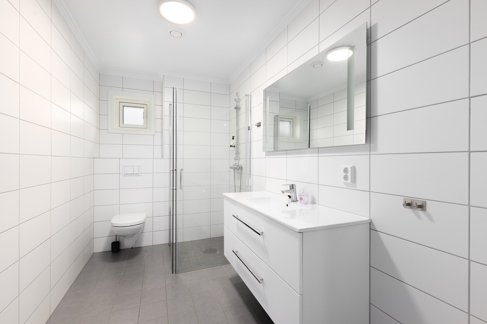 Flislagt bad med varmekabler i gulv. Badet er innredet med vegghengt toalett, dusjhjørne med glassdører og servantinnredning med skuffer og speil. Galleribilde