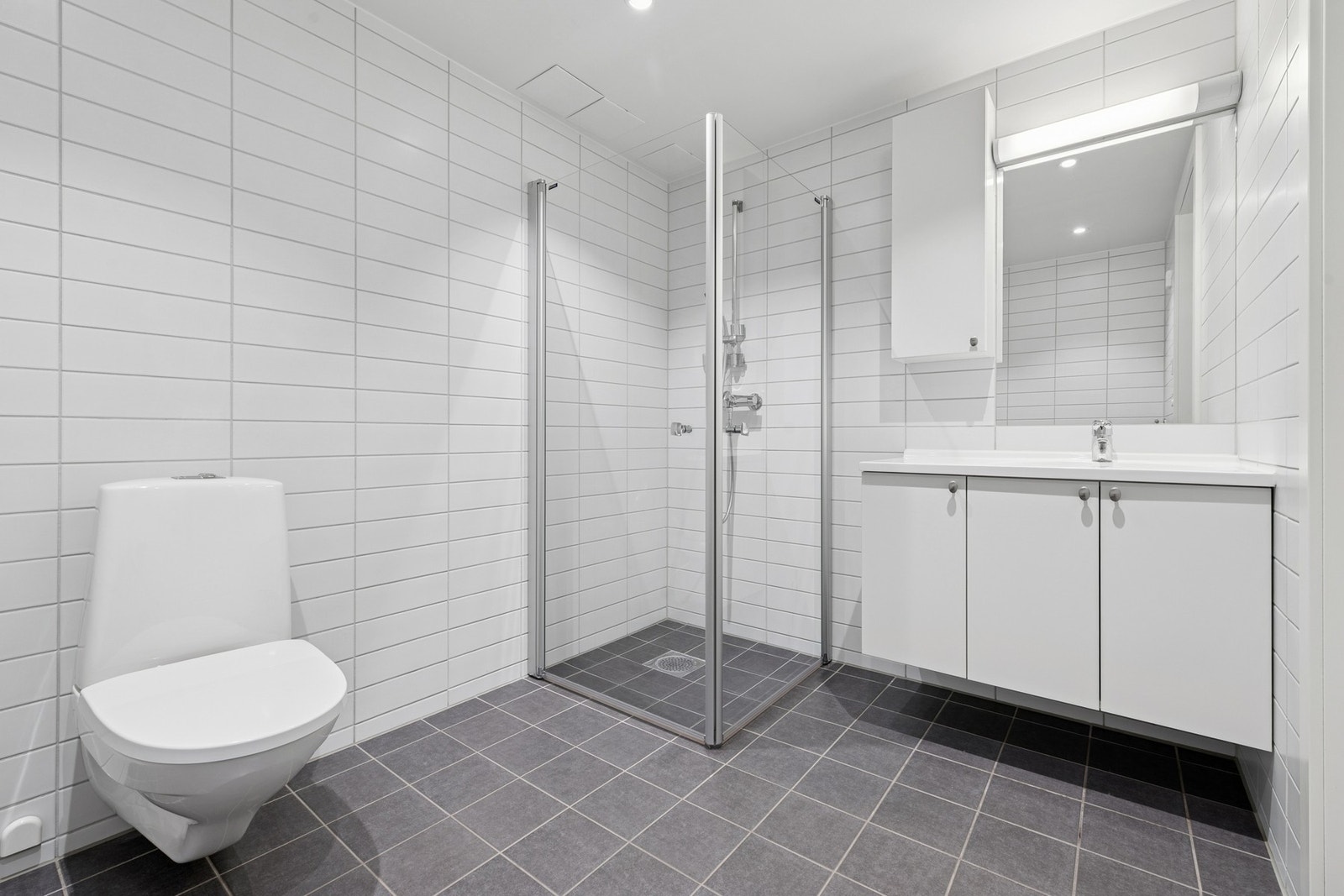 Lekkert og romslig bad fra byggeår med moderne, tidløst uttrykk. Rommet er helfliset med grå gulvfliser og hvite veggfliser, samt varme i gulv for økt komfort. Galleribilde