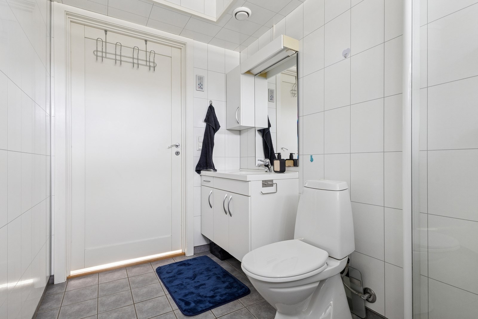 Badet er utstyrt med gulvmontert toalett, dusjhjørne med glassvegger, samt baderomsinnredning med servant og speil, samt opplegg for vaskemaskin. Galleribilde