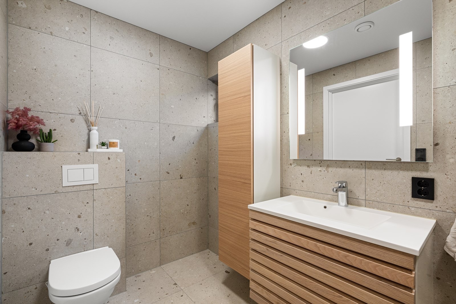 Badet er utstyrt med downlights i himling og varmekabler i gulv. Badet er innredet med vegghengt toalett, dusjhjørne, servant med underskap og speil med belysning. Galleribilde