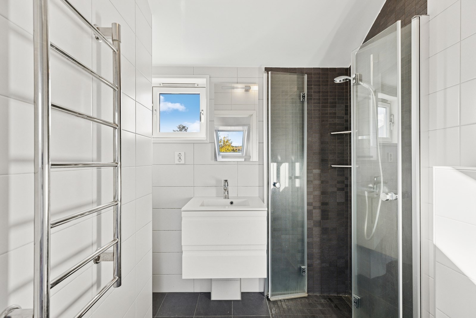 Badet er tidløst innredet med en kombinasjon av grå og hvite fliser. I gulvet har man også varmekabler for ekstra komfort. Badet er ellers utstyrt med vegghengt toalett, dusjhjørne, elektrisk håndkletørker og baderomsinnredning med nedfelt servant. Galleribilde