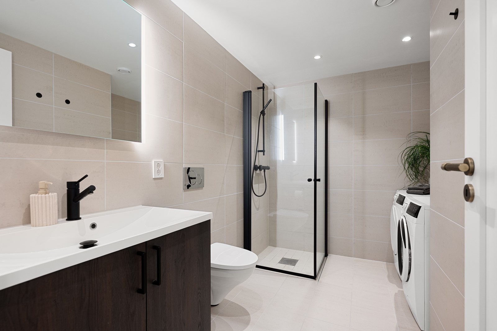 Boligens andre bad er kombinert med et praktisk vaskerom og har adkomst fra gangen i underetasjen. Rommet følger samme stil og materialvalg som badet ved hovedsoverommet. Galleribilde