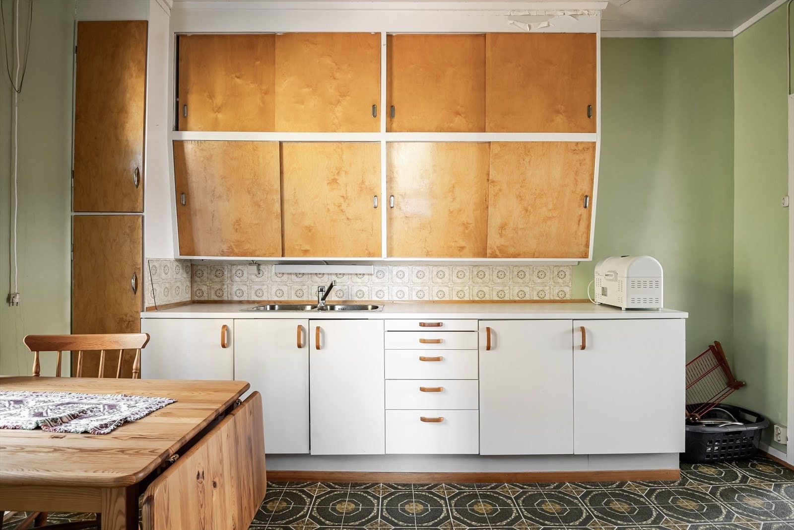 Eldre kjøkkeninnredning med glatte fronter, laminert benkeplate med nedfelt oppvaskkum og vinylbelegg i benkeryggen. Montert håndvask på vegg. Galleribilde