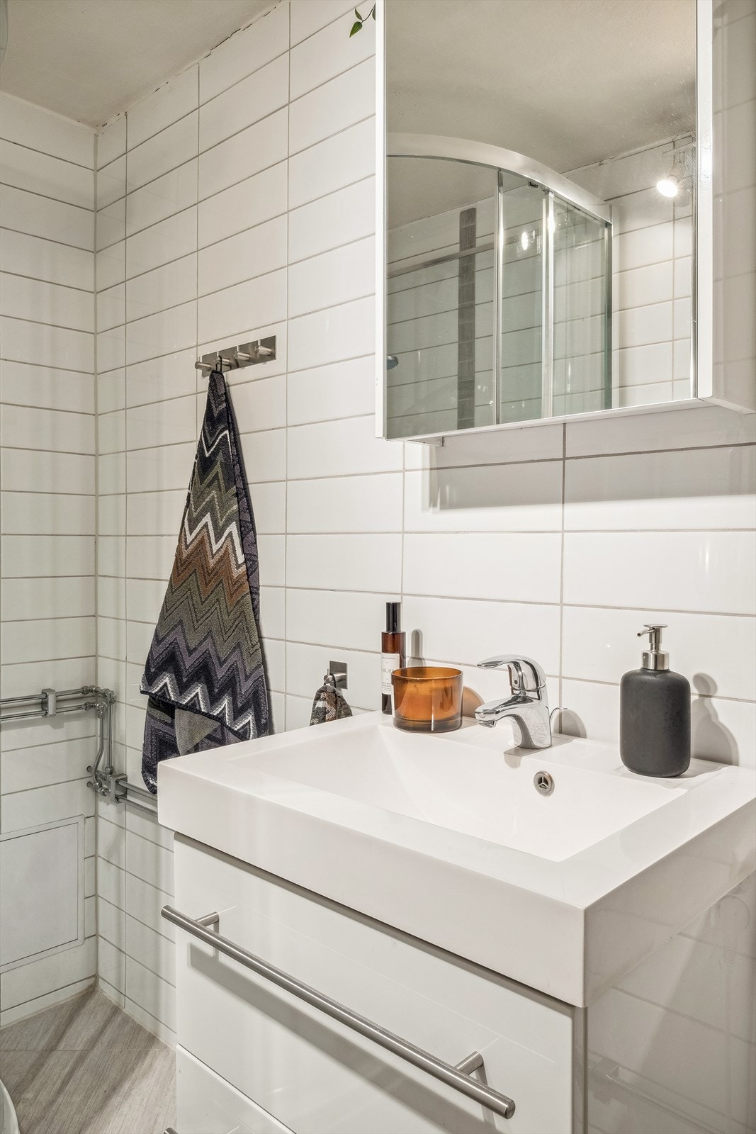 Badet er innredet med heldekkende servant i hvit skuffeinnredning, speilskap, dusjkabinett og gulvmontert toalett. Det er etablert elektrisk gulvvarme i badegulvet. Det er også naturlig ventilering. Galleribilde