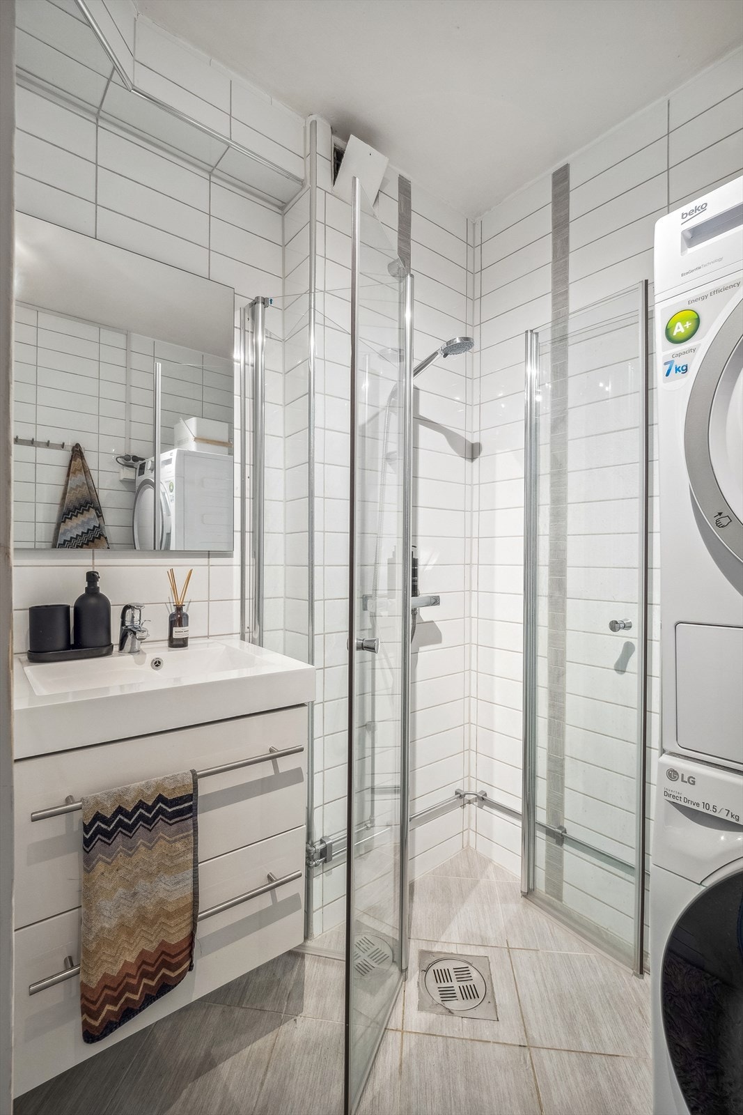 Bad nr 2 har hvite fliser på veggene og sand/gråfargede fliser på gulvet. Badet er innredet med heldekkende servant i hvit skuffeinnredning, speil på vegg, dusjvegger med dusjarmatur montert på vegg og opplegg for vaskemaskin. Galleribilde