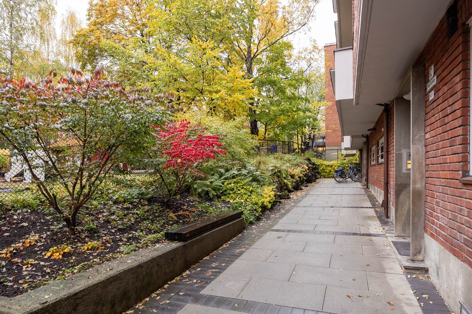 Bilde viser en utvendig gangvei med belegningsstein og frodig beplantning langs fasaden. Gangveien gir enkel tilgang til leilighetens inngangsparti. Galleribilde