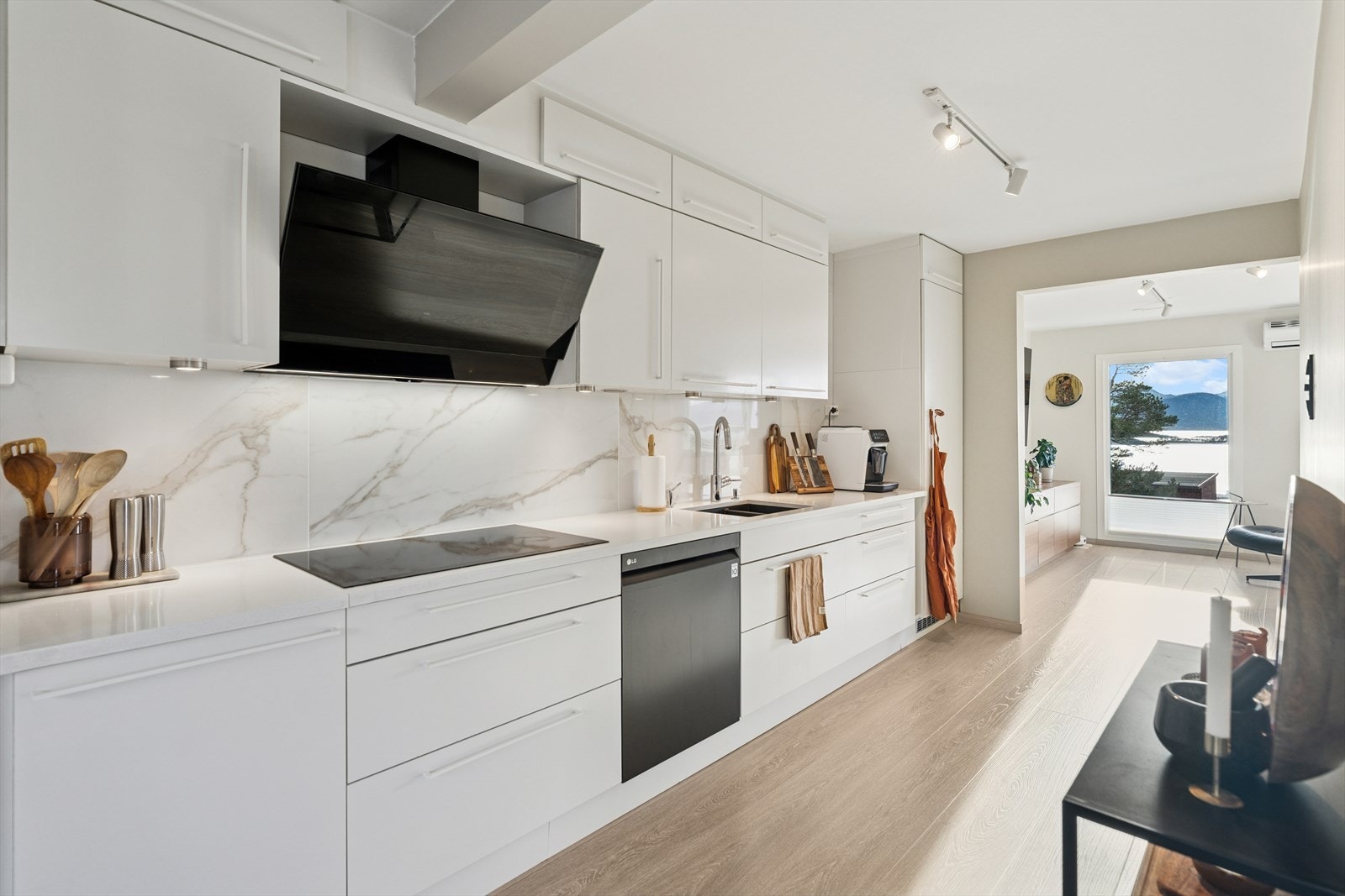 Lyst og pent HTH-kjøkken med glatte fronter, steinbenkeplate og flislagt vegg mellom benk og overskap. Laminatgulv og malte, slette overflater gir et moderne uttrykk. Galleribilde