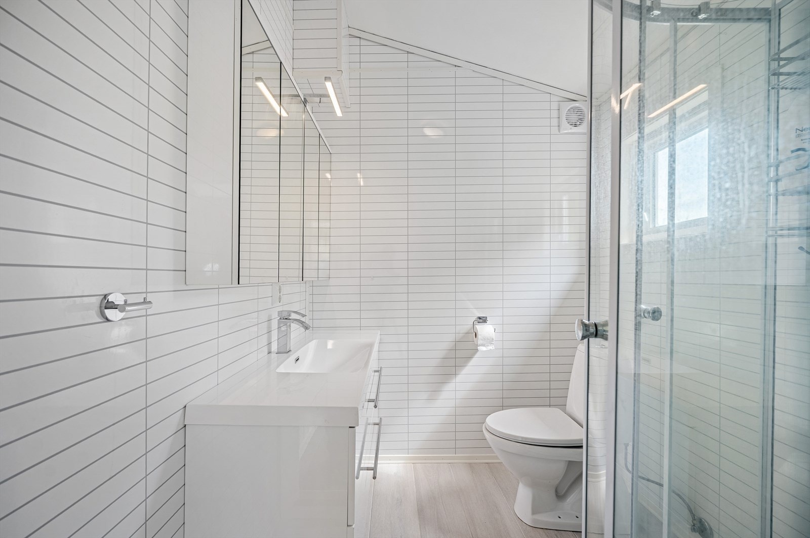 Badet er lyst med fliser på gulv og baderomspanel på vegger. Det har dusj, toalett og servant i baderomsinnredning. Galleribilde