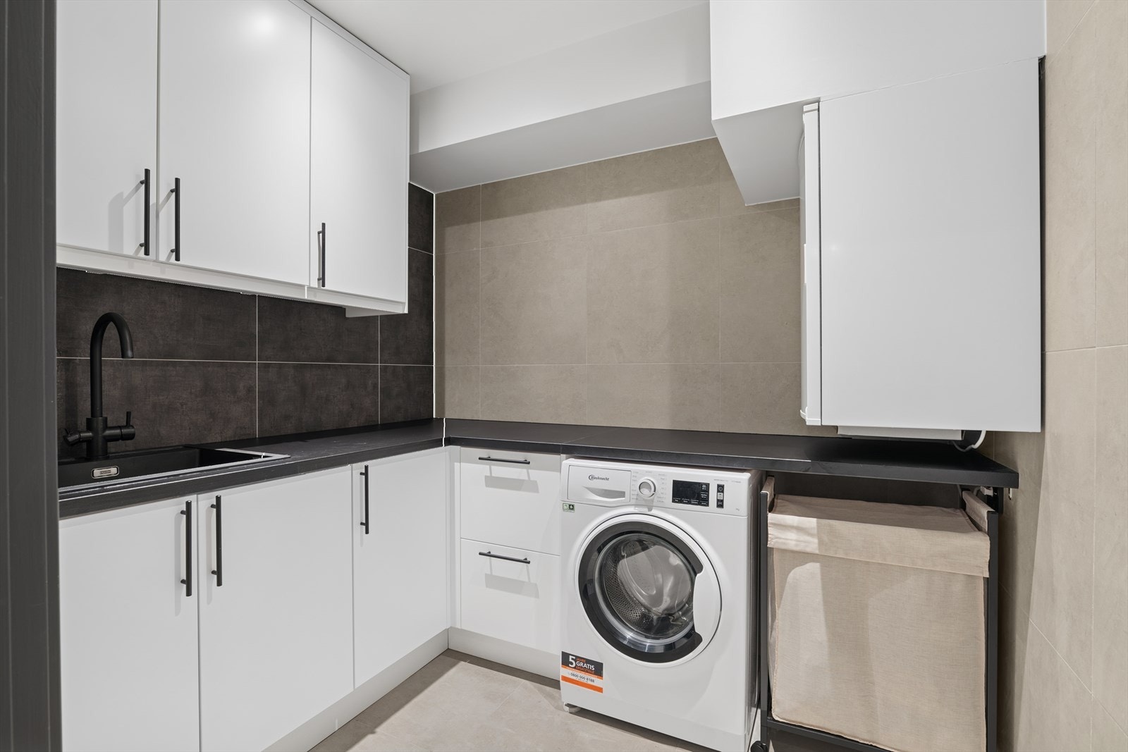 Fra badet er det videre inngang til et eget vaskerom via plassbesparende skyvedør som adskiller rommene pent fra hverandre. Rommet er praktisk innredet med opplegg for vaskemaskin, skapplass for oppbevaring, servant i sort utførelse og benkeplass. Galleribilde