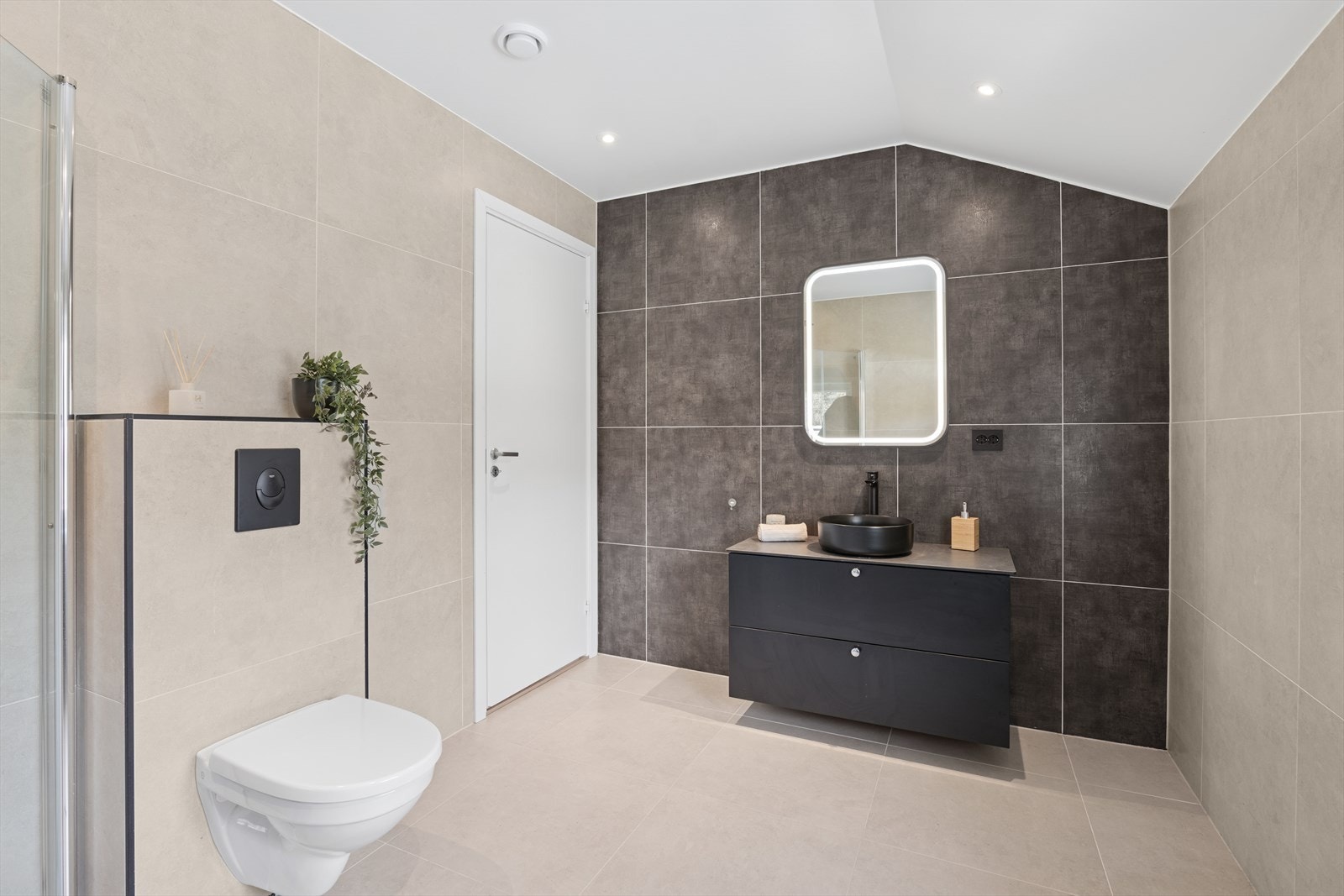 Badet er innredet med stor dusjnisje med glassdør, vegghengt toalett, baderomsinnredning med skuffeseksjon, bolleservant og belyst vegghengt speil. Det er vindu på vegg som sikrer rommet godt med daglys, samt fin spotbelysning i taket. Galleribilde