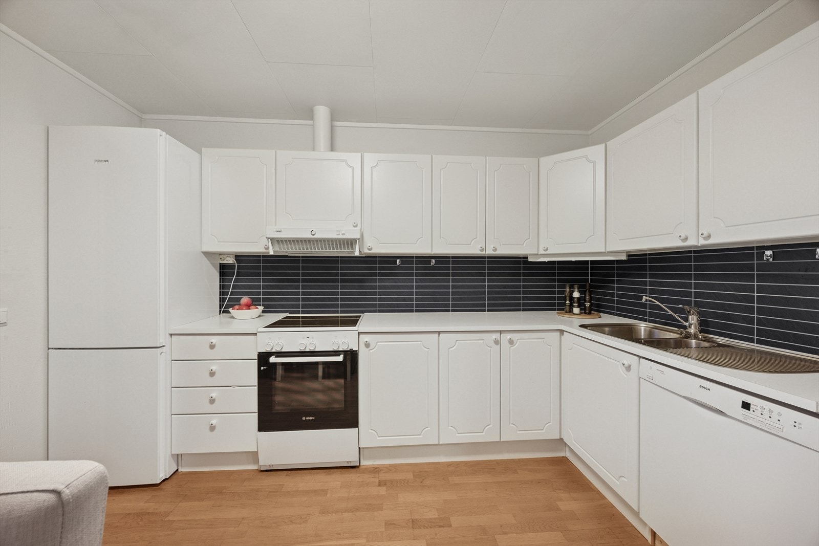 Her finner du en innredning med hvite profilerte fronter som gir et lyst og tidløst uttrykk. Den slitesterke laminat benkeplaten gir god arbeidsflate, og kjøkkenet er utstyrt med rikelig skapplass og benkeplass. Galleribilde