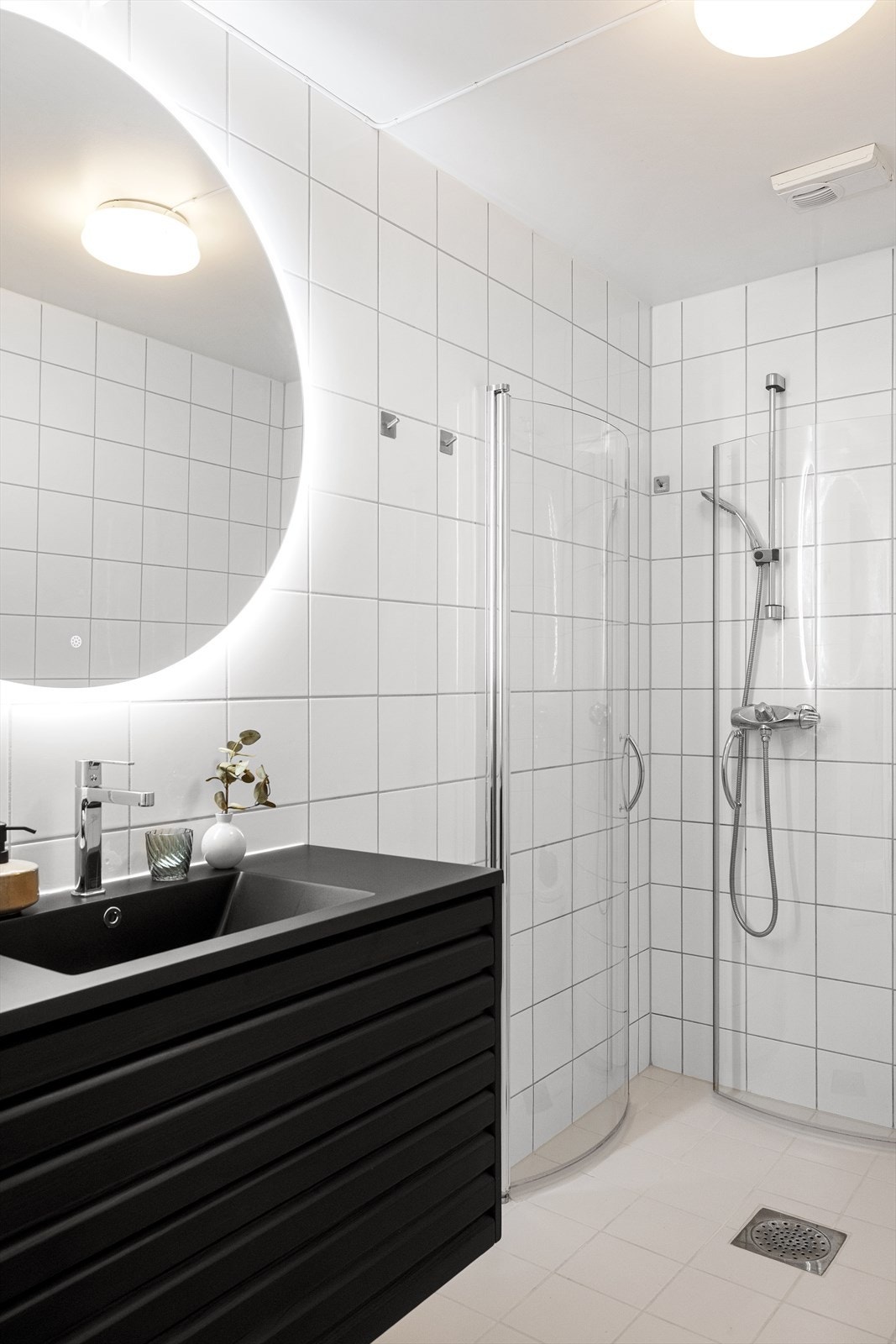 Baderommet har dusjnisje med glassdører og moderne armatur. Helhetlig uttrykk med mørk innredning og kontrast mot lyse fliser. Galleribilde
