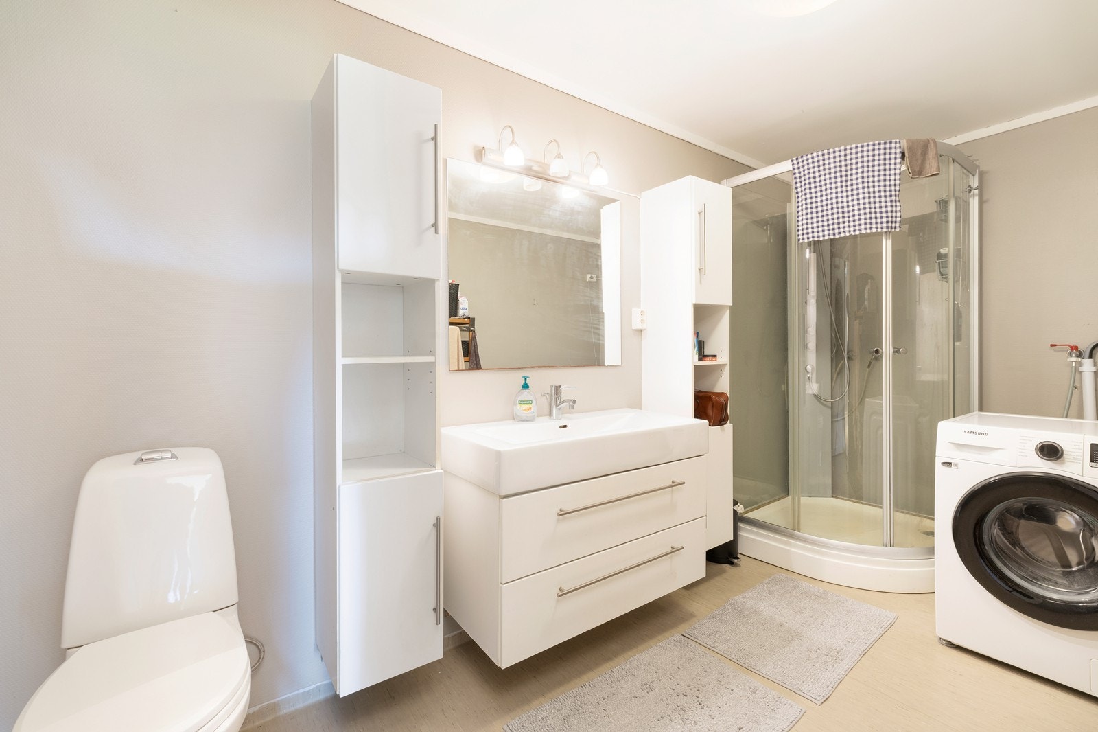 Bad med belegg på gulv. Badet har dusjkabinett, toalett, servant med underskuffer og opplegg til vaskemaskin. Galleribilde