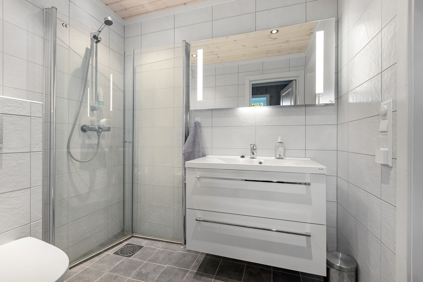 De lyse flisene og panel i himling skaper et rent og tidløst uttrykk. Pen baderomsinnredning med to skuffer og heldekkende servantplate. Galleribilde