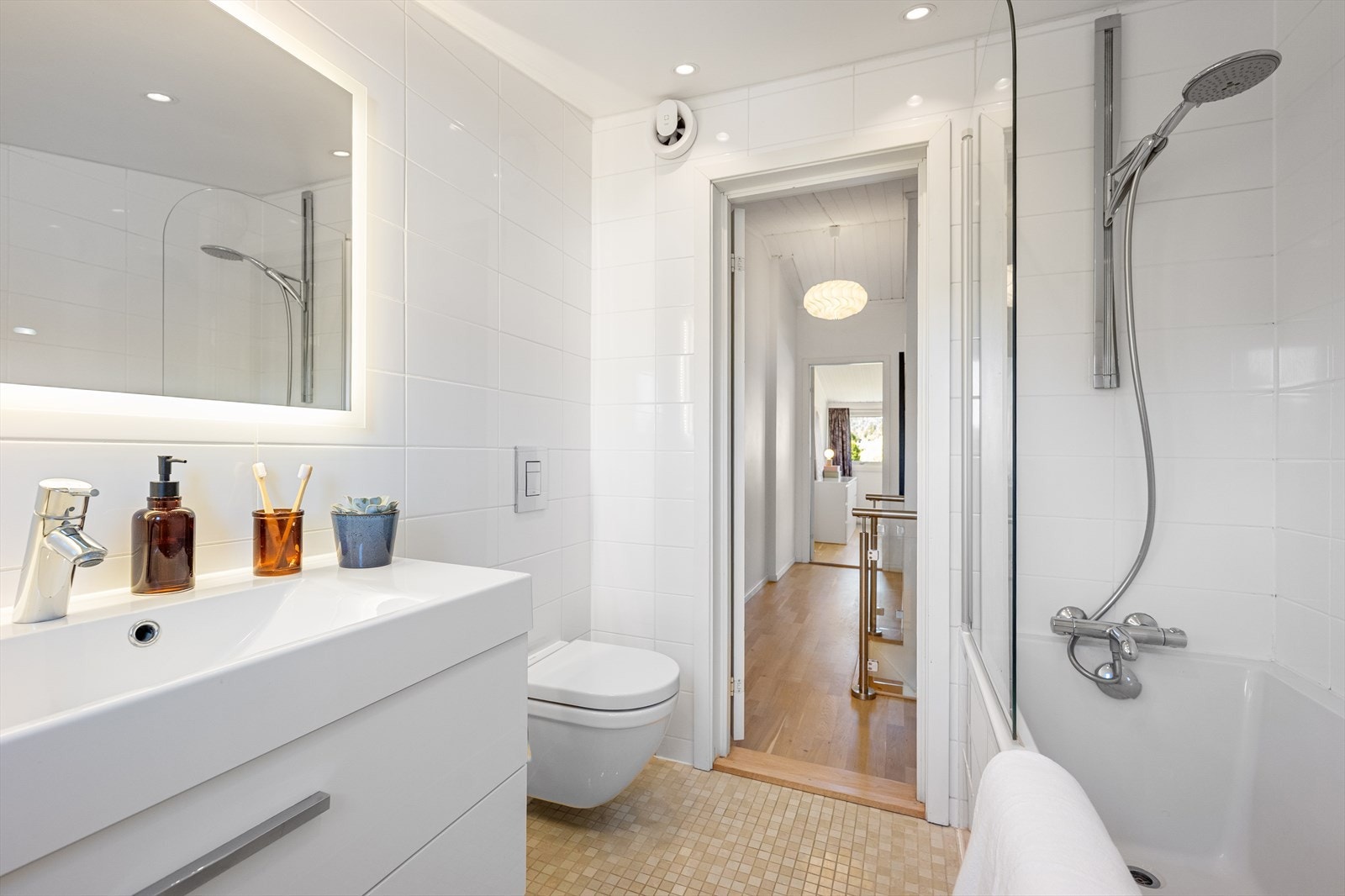 Badet er innredet med vegghengt toalett, servant i skuffeinnredning, speil med integrert belysning og badekar med dusj og dusjvegg. Galleribilde