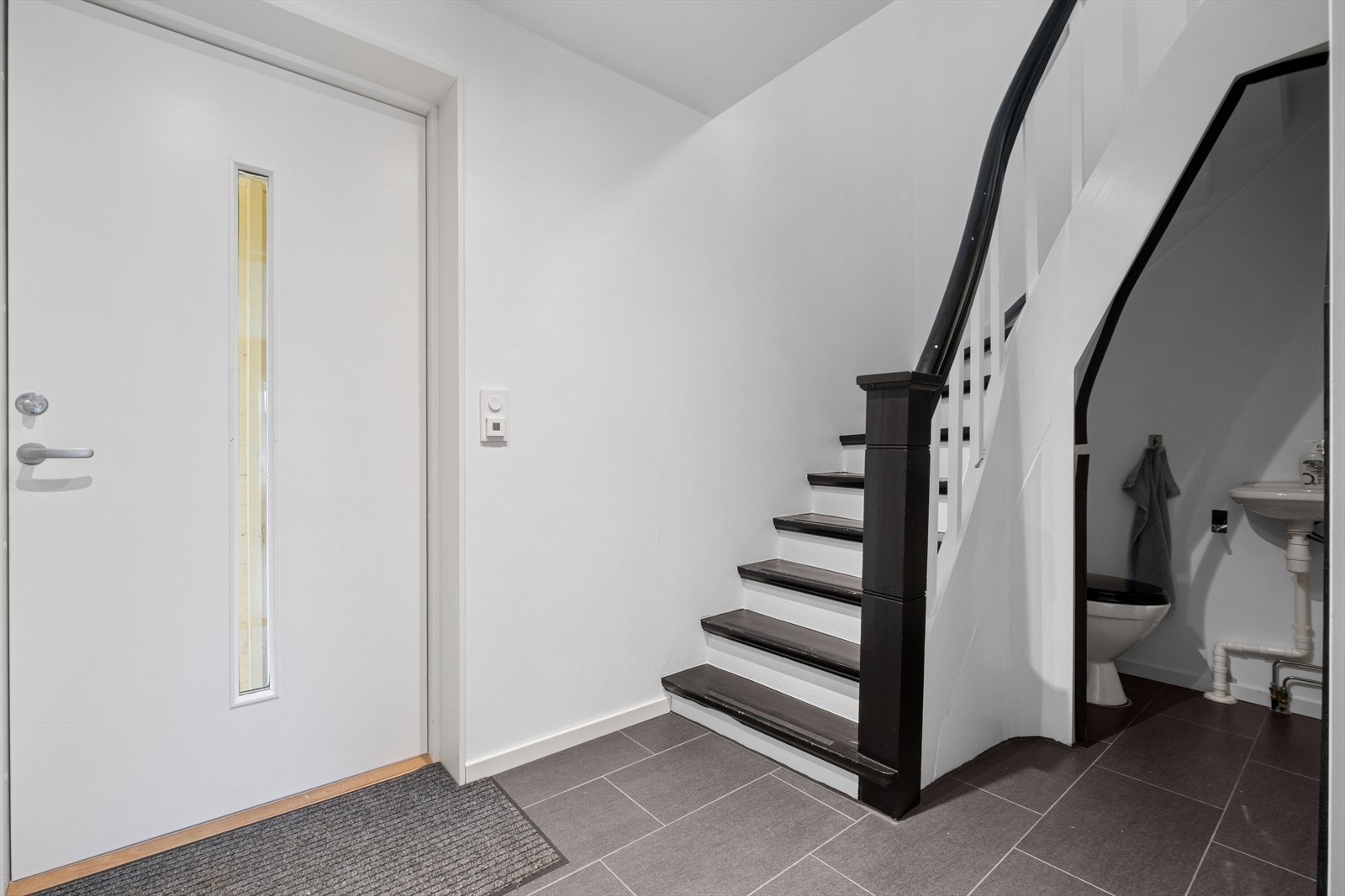 Entréen fremstår som lys og innbydende, med lyse vegger og store, grå gulvfliser som gir et moderne og praktisk uttrykk. Rommet har god plass til å henge fra seg yttertøy og sette fra seg sko. Galleribilde