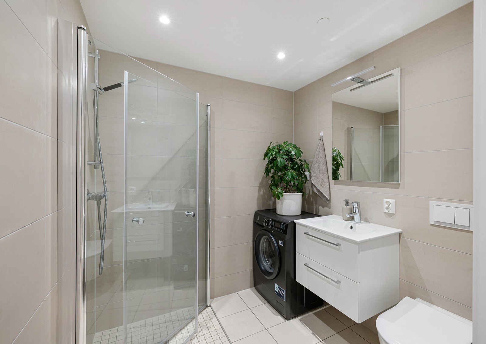 Badet har en stilren og tidløs utforming med lyse fliser på vegger og gulv. Det er opplegg for vaskemaskin, og gulvvarme gir ekstra komfort. Galleribilde