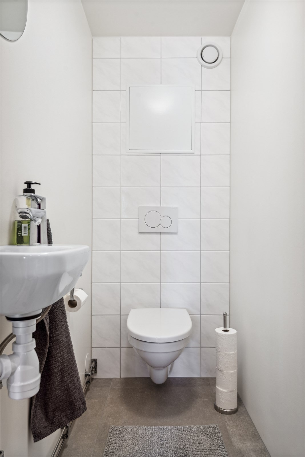 Toalettrommet har grå keramiske fliser på gulvet, lysmalt mur, plater og en vegg med hvite keramiske fliser på veggene. Toalettrommet er innredet med servant montert på vegg og veggmontert toalett. Galleribilde