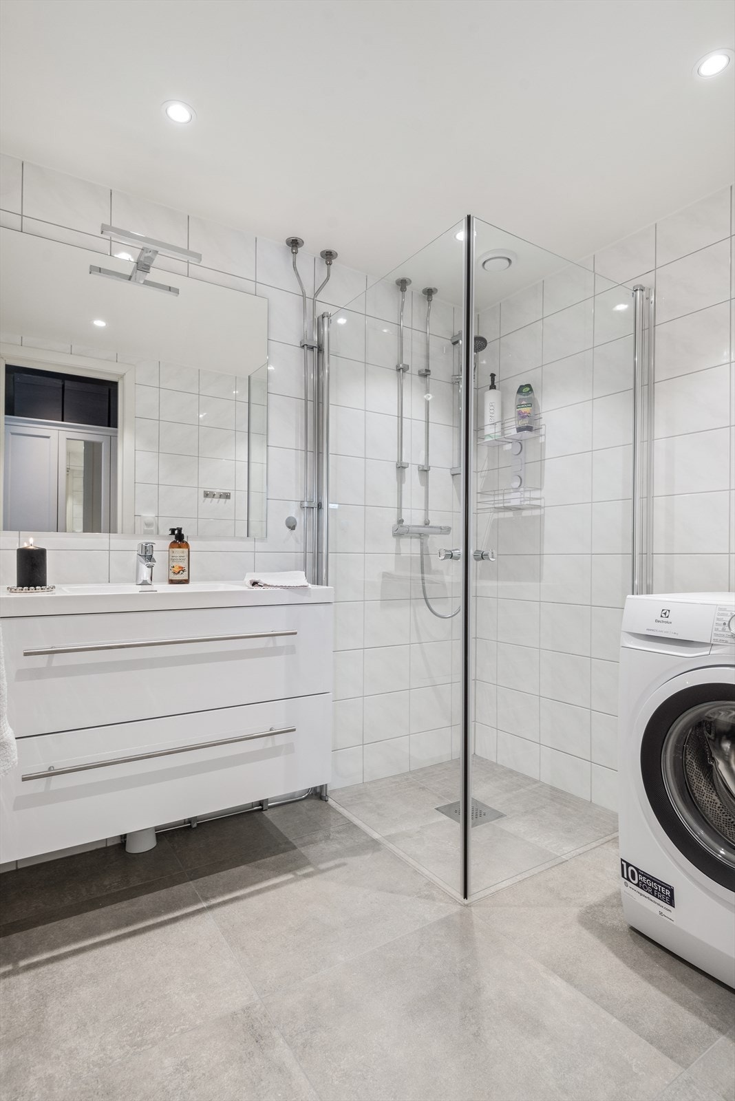 Badet har hvite keramiske fliser på veggene og grå fliser på gulvet. Badet er innredet med heldekkende servant i hvit skuffeinnredning, speil, dusjhjørne med dører og dusjarmatur montert på vegg og opplegg for vaskemaskin. Galleribilde