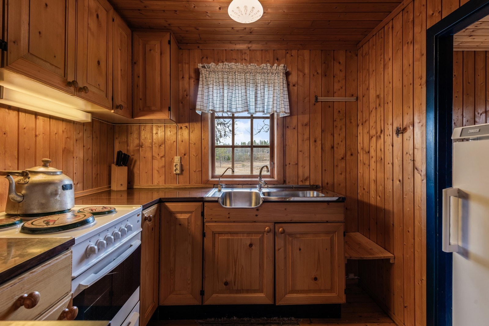 Kjøkkeninnredningen består av profilert treverk med både under- og overskap, benkeplate i laminat og dobbel oppvaskkum i rustfritt stål. Til høyere for bildet er det tilgang til hyttens første soverom. Galleribilde