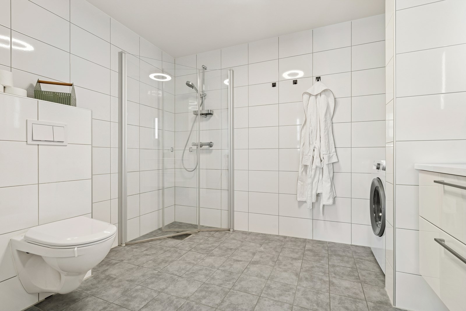 Baderommet er innredet med vegghengt toalett, servant, Opplegg for vaskemaskin og åpen dusjsone med dusjgarnityr på vegg Galleribilde