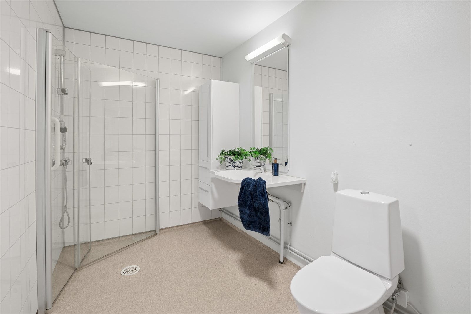 Badet er lyst og pent og har belegg på gulv med varmekabel. Veggene har tapet og baderomsplater, og himlingen har malt betong. Rommet er innredet med klosett, servant og dusjhjørne med svingbare dører. Galleribilde