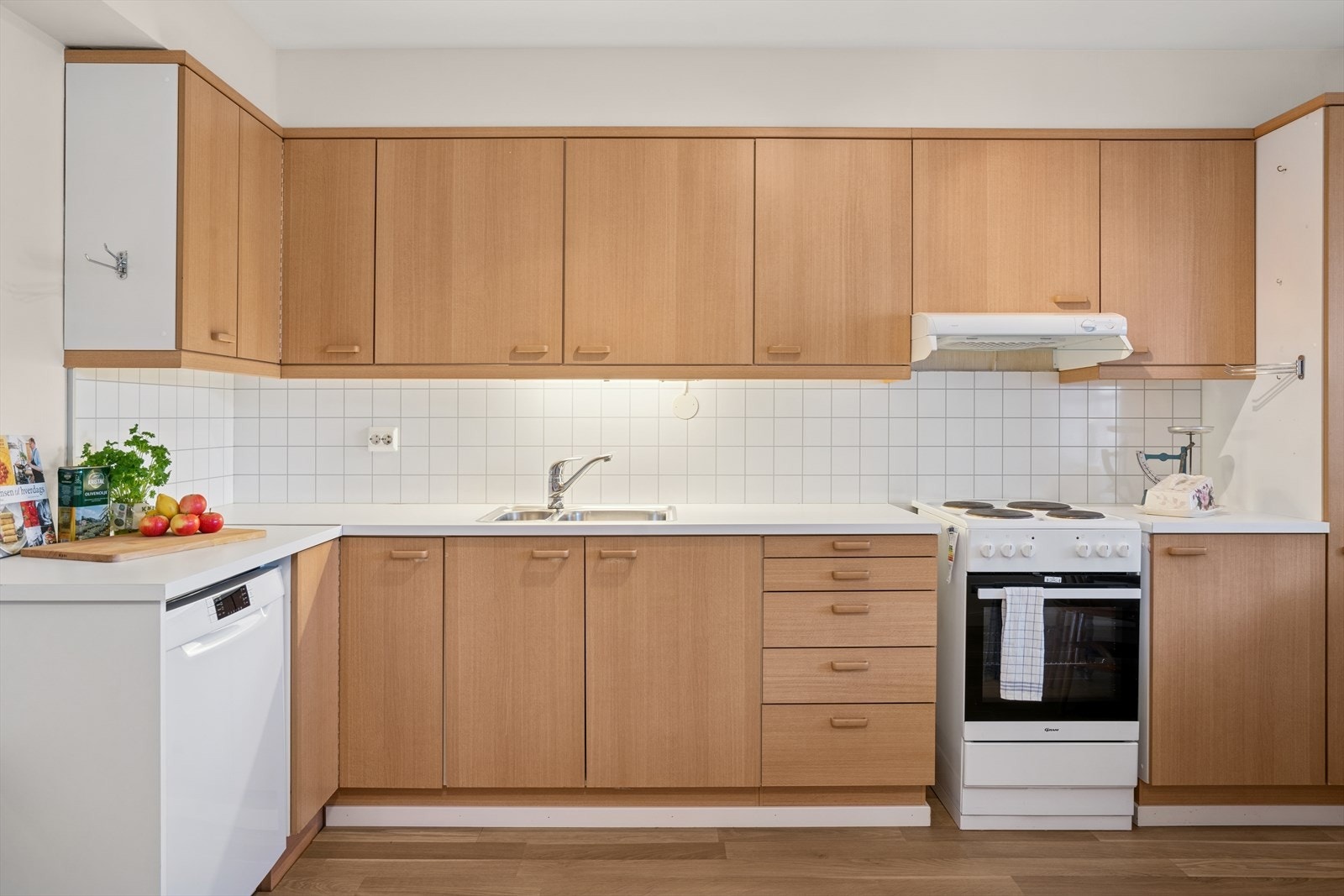 Kjøkkenventilator over kokeplass med avtrekk via mekanisk anlegg. Det er medfølgende frittstående eldre komfyr og oppvaskmaskin. Galleribilde