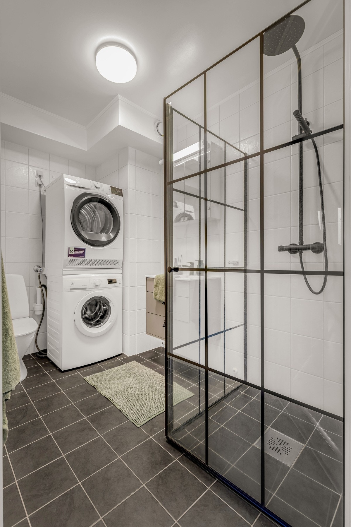 Badet har et tidløst design med fliser i gulv og vegger, servant med skuffer under, dusjvegger, toalett og opplegg for vaskemaskin. Galleribilde