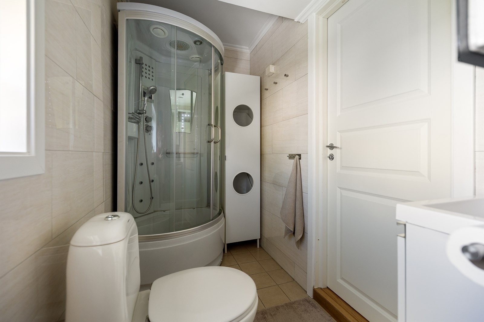 Taket er utført med malte plater og downlights, noe som tilfører rommet en moderne og behagelig belysning. Rommet er innredet med dusjkabinett med kar og dyser, toalett, servantskap med god oppbevaringsplass, speil og et praktisk høyskap. Galleribilde