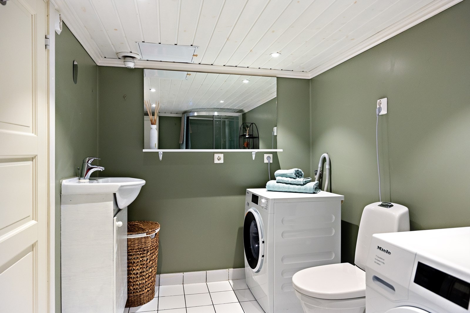 Praktisk baderom i kjeller. Her er både gulv og vegger flislagt, med oppkantflis som gir en helhetlig og slitesterk utførelse. Veggene kombineres med malte plater, mens taket har malt panel med innfelte downlights som gir et rent og moderne uttrykk. Galleribilde