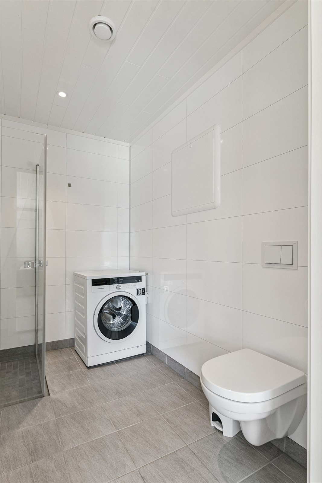 Bad med opplegg for vaskemaskin i underetasje. Flis på golv med varmekablar og baderomsplater på vegg.
Veggmontert klosett, dusj med hengsla glasdører, heilstøypt vask innredning og opplegg til vaskemaskin. Galleribilde