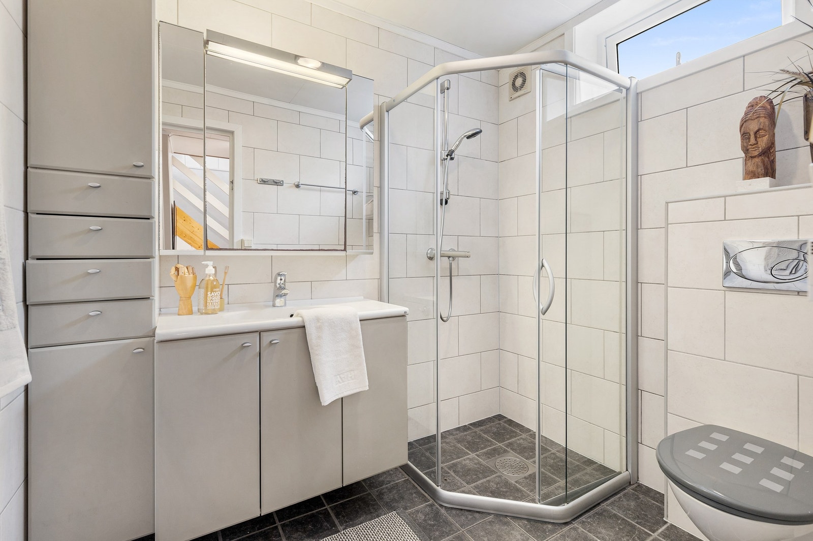 Flislagt gulv med varme og flislagte vegger. Himling med hvite himlingsplater. Vegghengt servantskap. Ovenpåliggende servant med ett-greps armatur. Speilskap med belysning på vegg over servant. Galleribilde