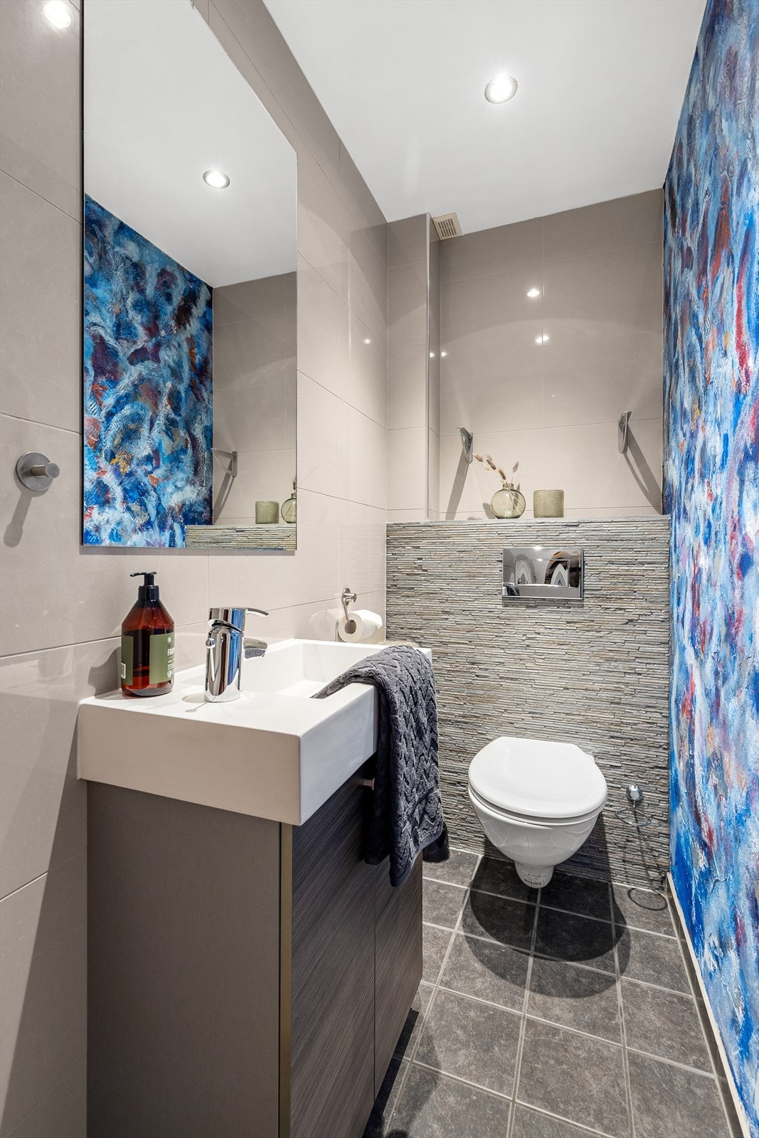 Flislagt gulv med varme. Vegger med fliser/malt mur. Nedsenket himling med downlights. Vegghengt servantskap. Ovenpåliggende servant med ett-greps armatur. Speil på vegg over servant. Vegghengt toalett. Naturlig avtrekksventil i himling. Galleribilde