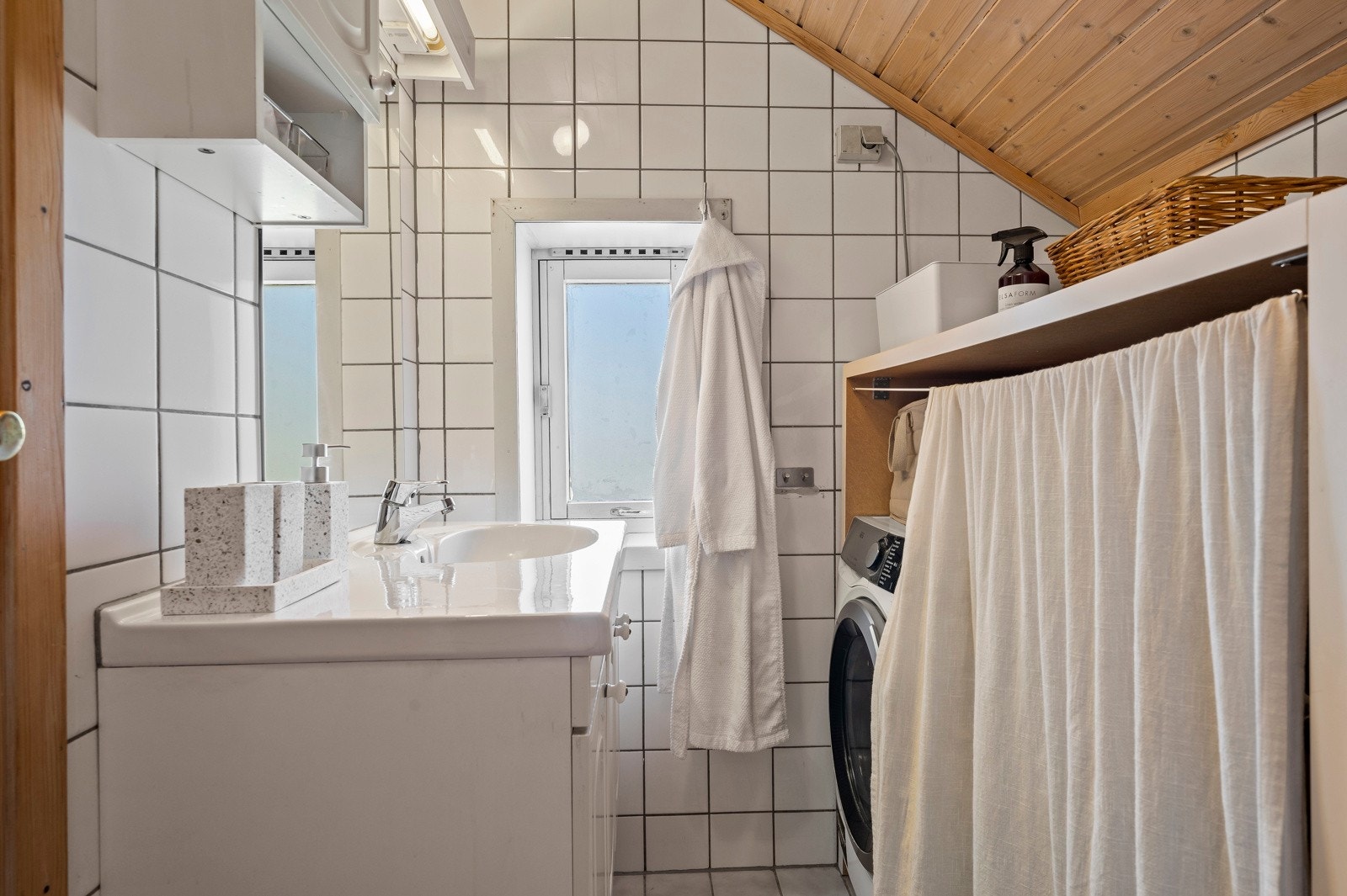 Et effektivt innredet bad med lyse overflater og plassbesparende løsninger som inkluderer dusjkabinett, toalett, vaskemaskin og servant Galleribilde