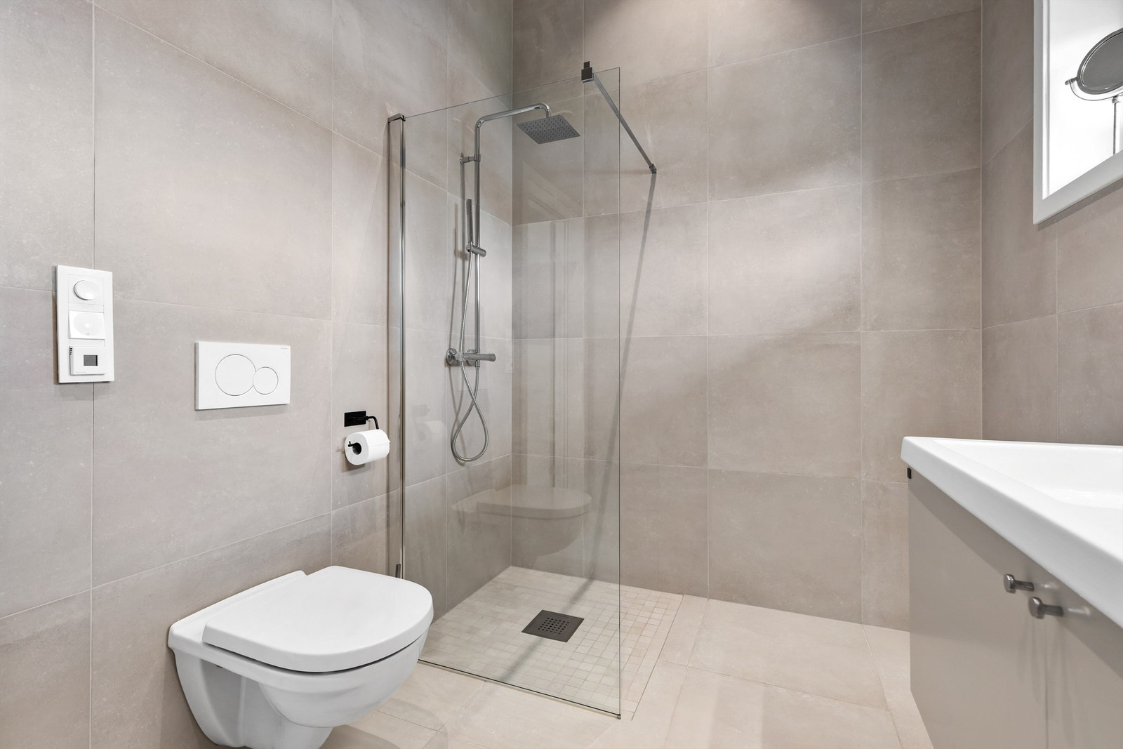 Ovenpåliggende dobbel servant med ett-greps armaturer. Speil og belysning samt stikkontakt på vegg over servant. Dusjsone med delvegg av glass. Dusjarmatur tilkoblet hånddusj og regndusj. Vegghengt toalett. Galleribilde