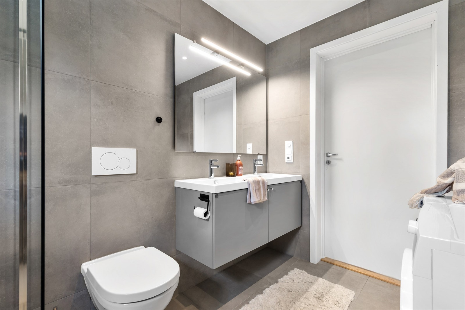 Baderom fra byggeår. Flislagt gulv med varme og flislagte vegger. Nedsenket himling med downlights. Vegghengt servantskap. Ovenpåliggende dobbel servant med ett-greps armaturer. Speil og belysning samt stikkontakt på vegg over servant. Galleribilde