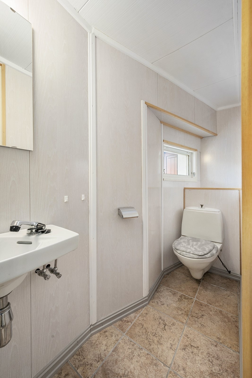 Toalettrom med fliser på gulv og baderomsplater på vegg. Rommet er innredet med servant, speil, vegglampe og toalett. Galleribilde