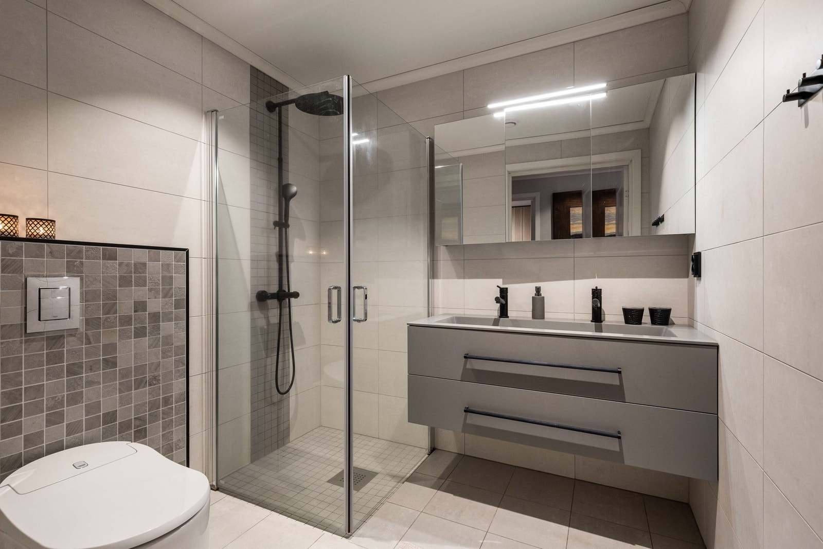 Badet i andre etasje - fliser på både gulv og vegger, varmekabler og moderne innredning i grå utførelse med skuffer, heldekkende servant og doble blandebatterier i sort. Galleribilde