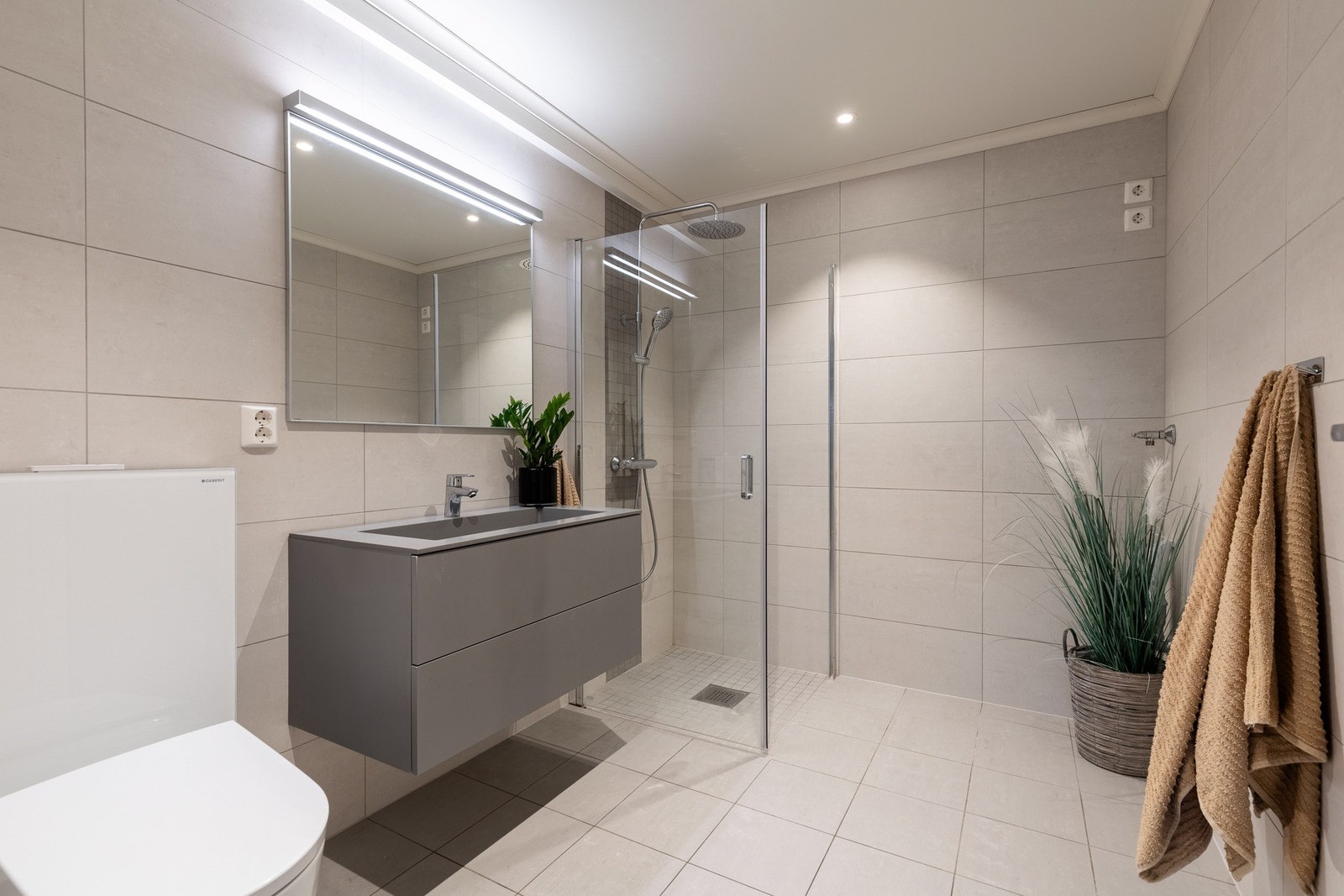 Badet i første etasje - fliser på gulv og vegger, varmekabler og innredning i grått. Her er det servant med skuffer, stort speil med belysning, dusjhjørne med glassdører, regnfallsdusj og toalett. Galleribilde