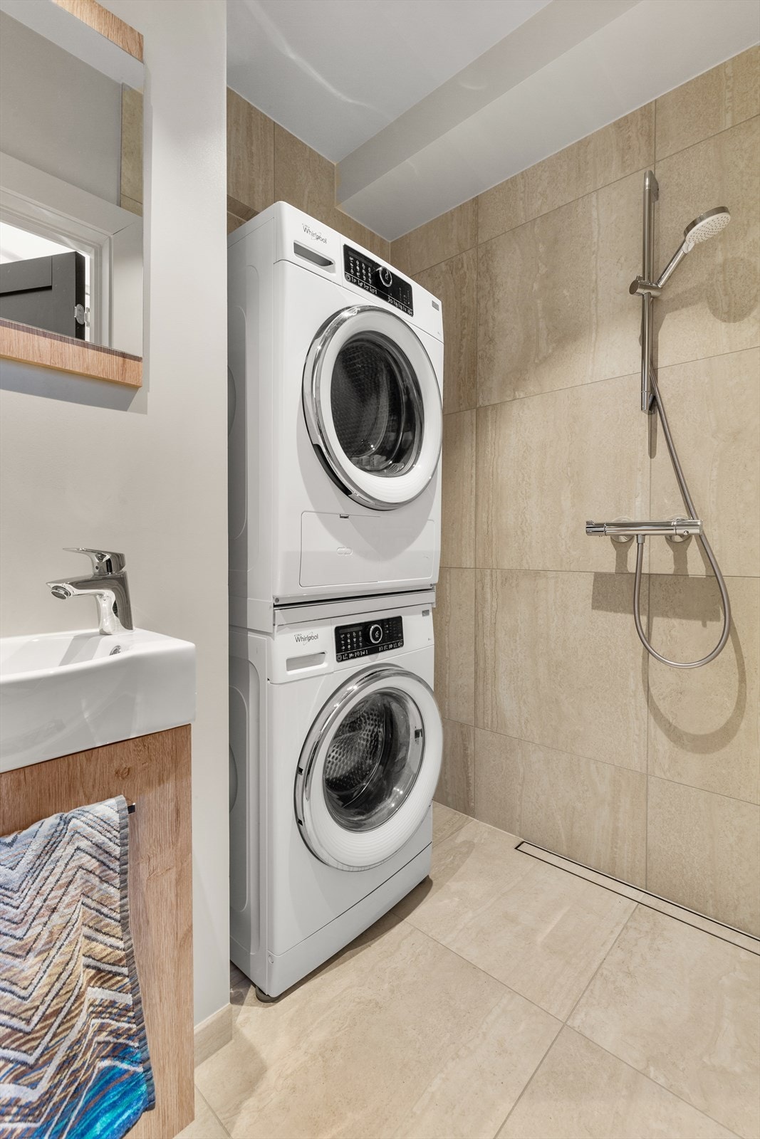 Lyst flislagt baderom med dusjnisje, servant med speil, vegghengt wc og opplegg for vaskemaskin og tørketrommel. Galleribilde