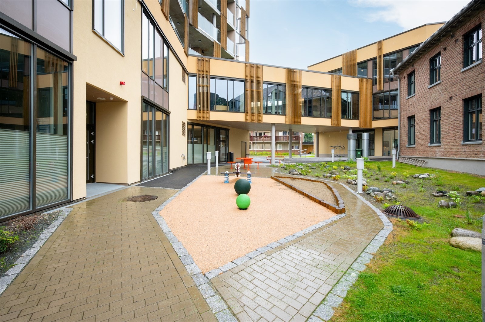 Inngangspartiet fra bakkeplan er pent opparbeidet med lekeapparater, grøntområde, sittebenker og belegningsstein. Galleribilde